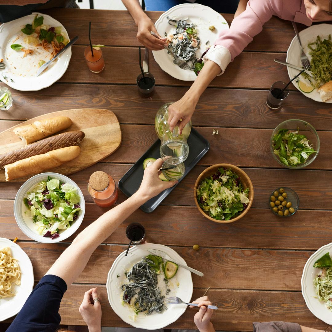 Top view of a wooden dining table with various dishes, including salads, pasta, bread, and drinks, as people reach for food and drinks, suggesting a communal & healthy meal setting